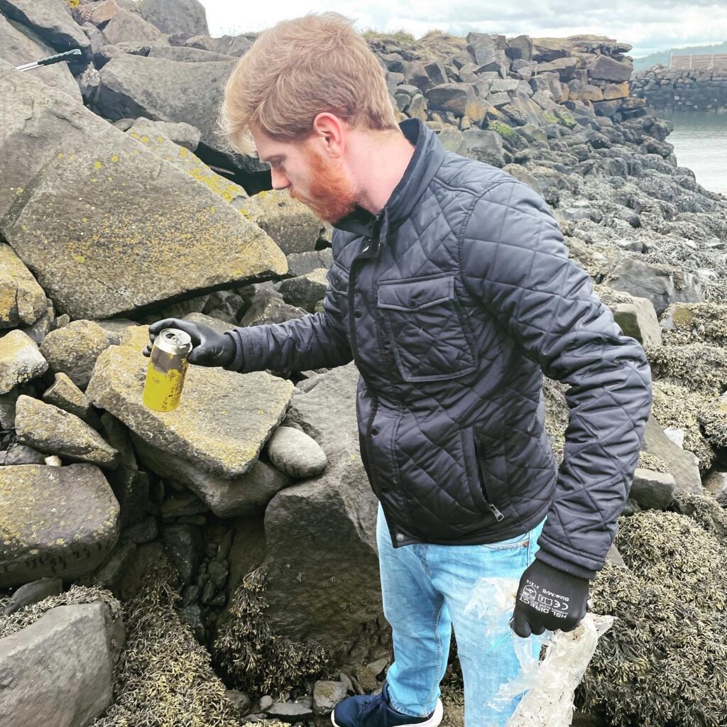 Volunteer picking up beer can on beach filled with sludge and dead small sea animals.